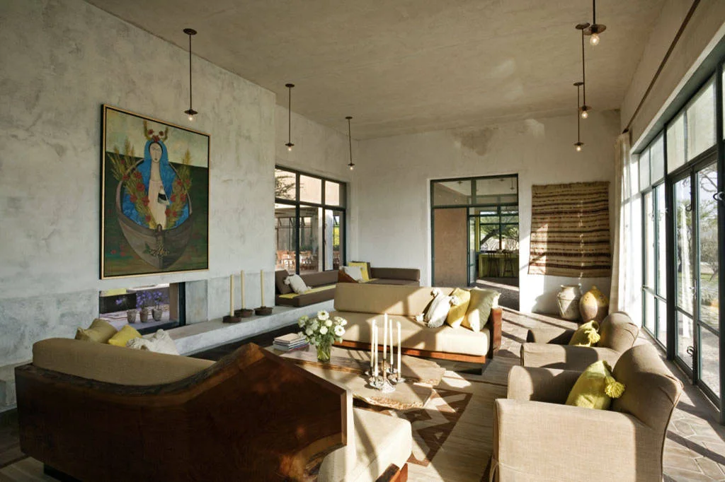 Neutral living room with wood-backed seating, yellow cushions, a colorful painting and a striped wall textile beside large windows.