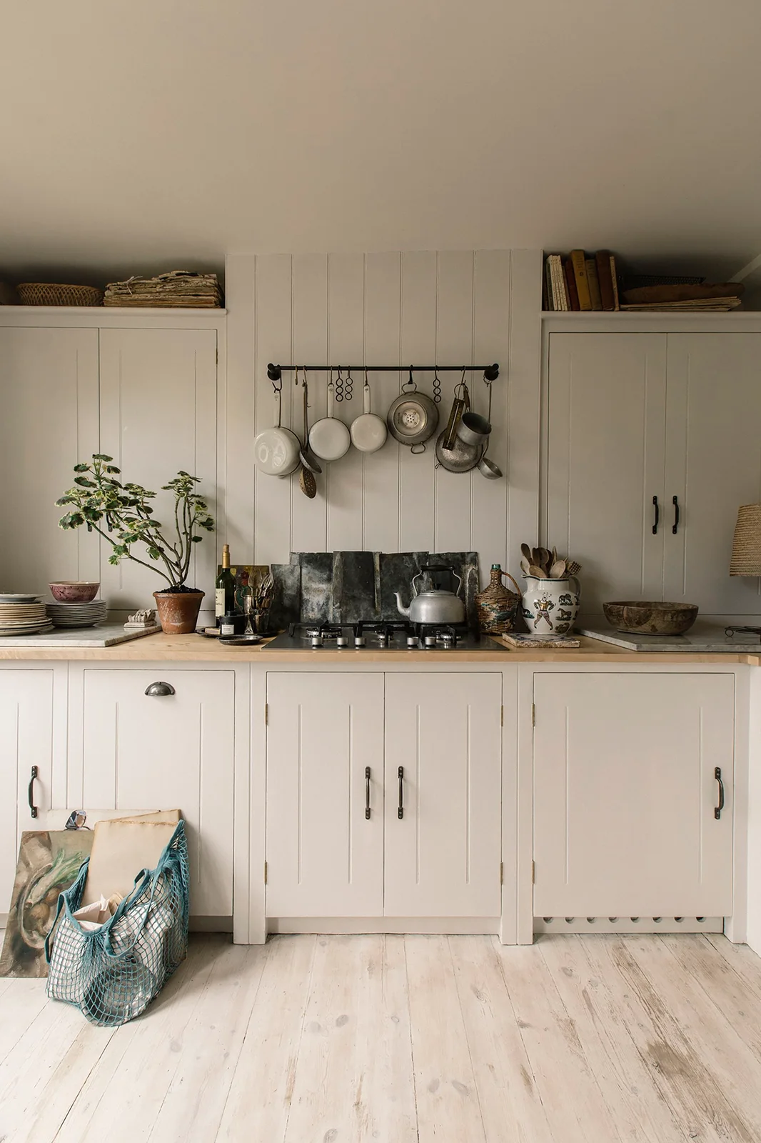 Creamy framed cupboards flank a pale boarded cooking wall with a dark rail and hanging pans.