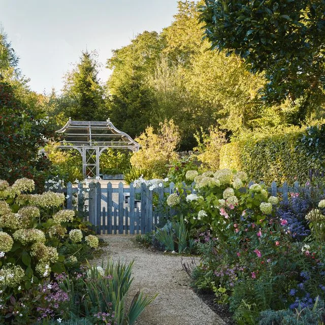 A curved gravel path leads through flowering borders to a blue picket gate and white arch