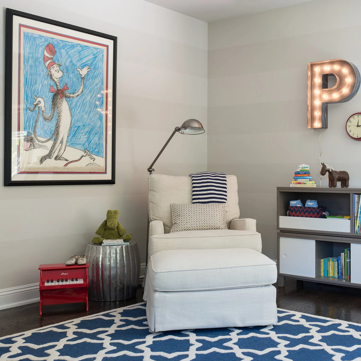 Pale nursery chair and ottoman beneath a large storybook print beside a metal-colored side table
