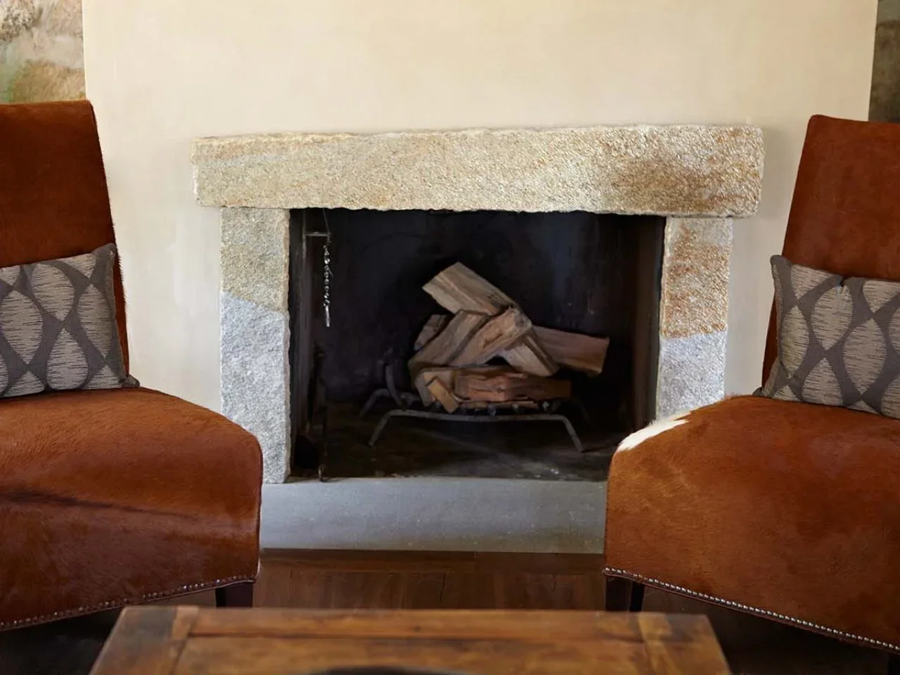 A rough pale stone header and uprights frame a dark fireplace between two rust-colored upholstered chairs.