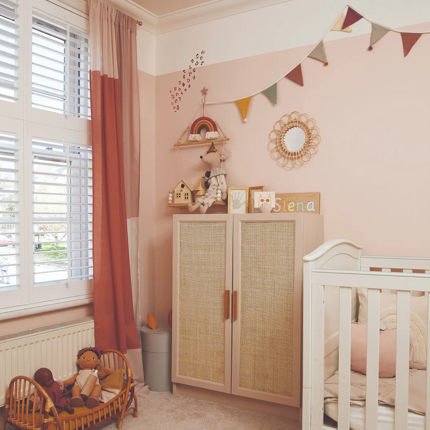 Pink nursery with white crib, woven-front cupboard, round decorative mirror, smaller wall pieces and pale window shutters.