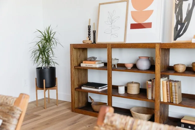A low open wood bookcase holds books, bowls and baskets beneath leaning framed prints, with a raised black plant pot at one end.
