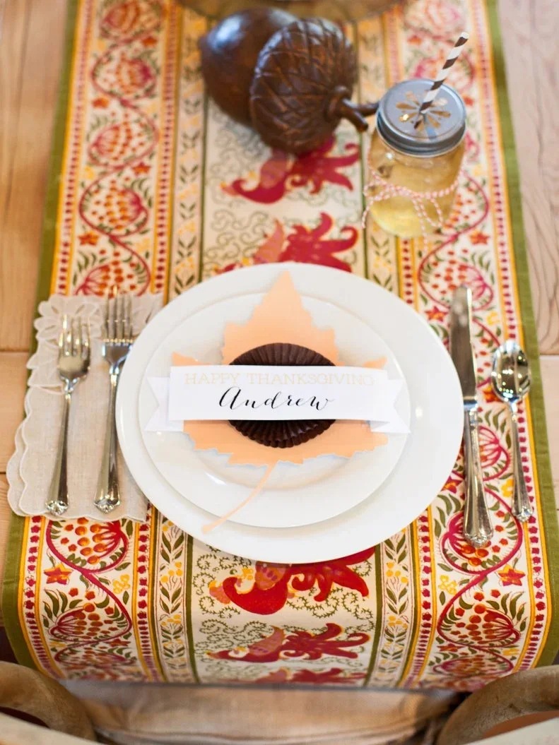 A name strip lies across a folded brown paper medallion and pale orange leaf shape on a white plate above patterned cloth