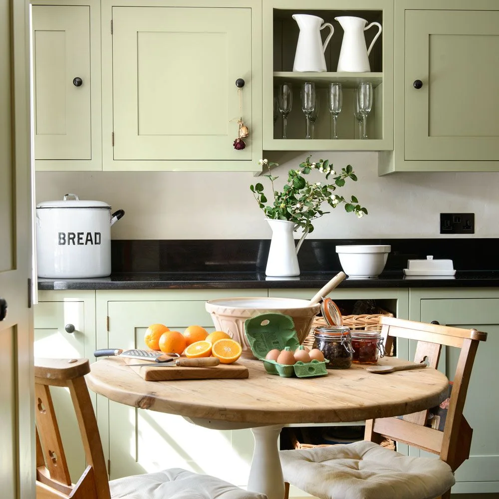 Pale green cupboards and an open glassware shelf sit behind a round wooden table beneath a dark counter