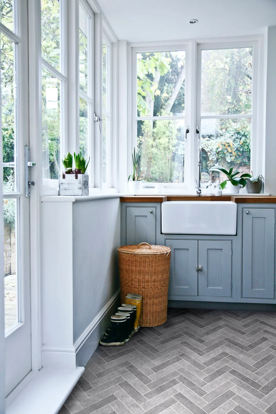 A white sink and pale blue framed cupboards sit under broad windows beside a wood counter, herringbone-patterned floor and a tall woven basket