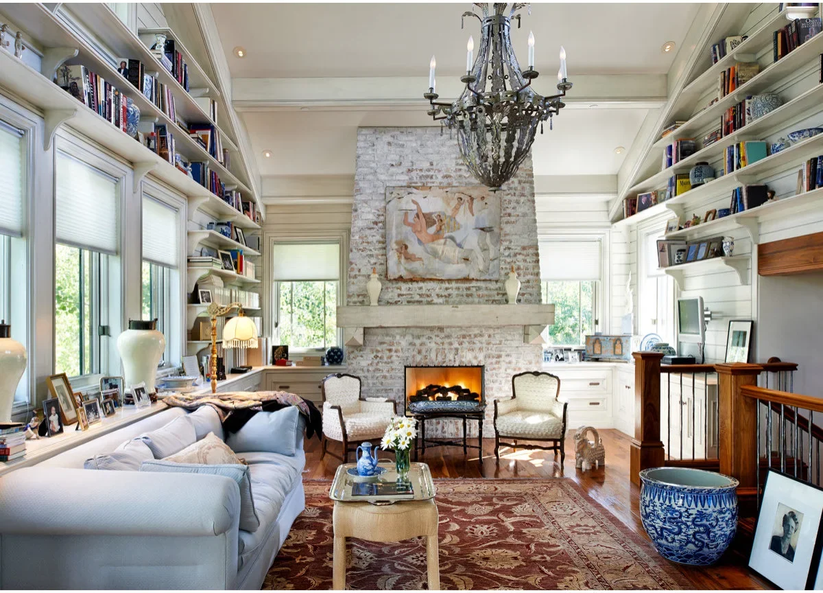 A tall pale, unevenly colored brick fireplace stands in a room with high pale bookshelves, a light sofa and a patterned red rug.