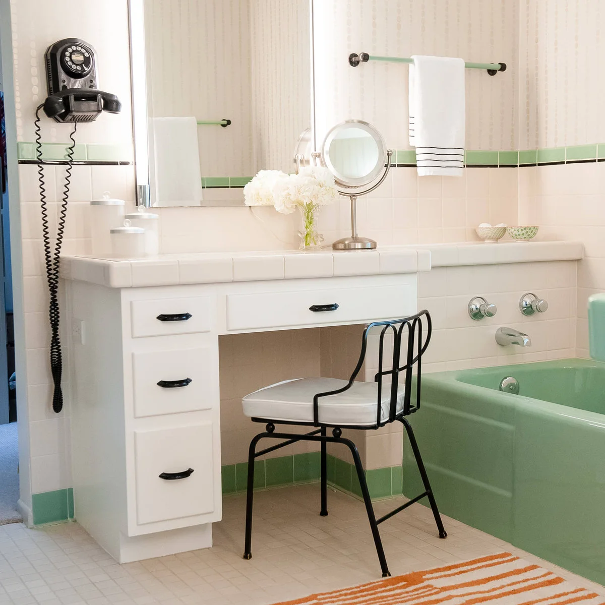 A white dressing table with black handles and a black metal chair sits beside a mint-green bath and green tile border.