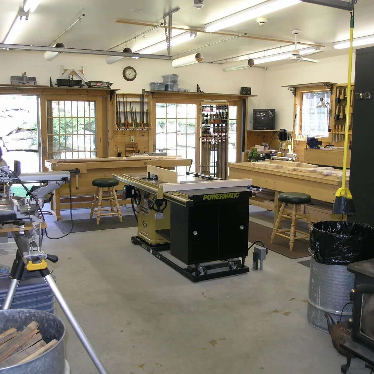 Woodworking shop with a central table saw, wooden assembly benches by windows and clamps hanging vertically.