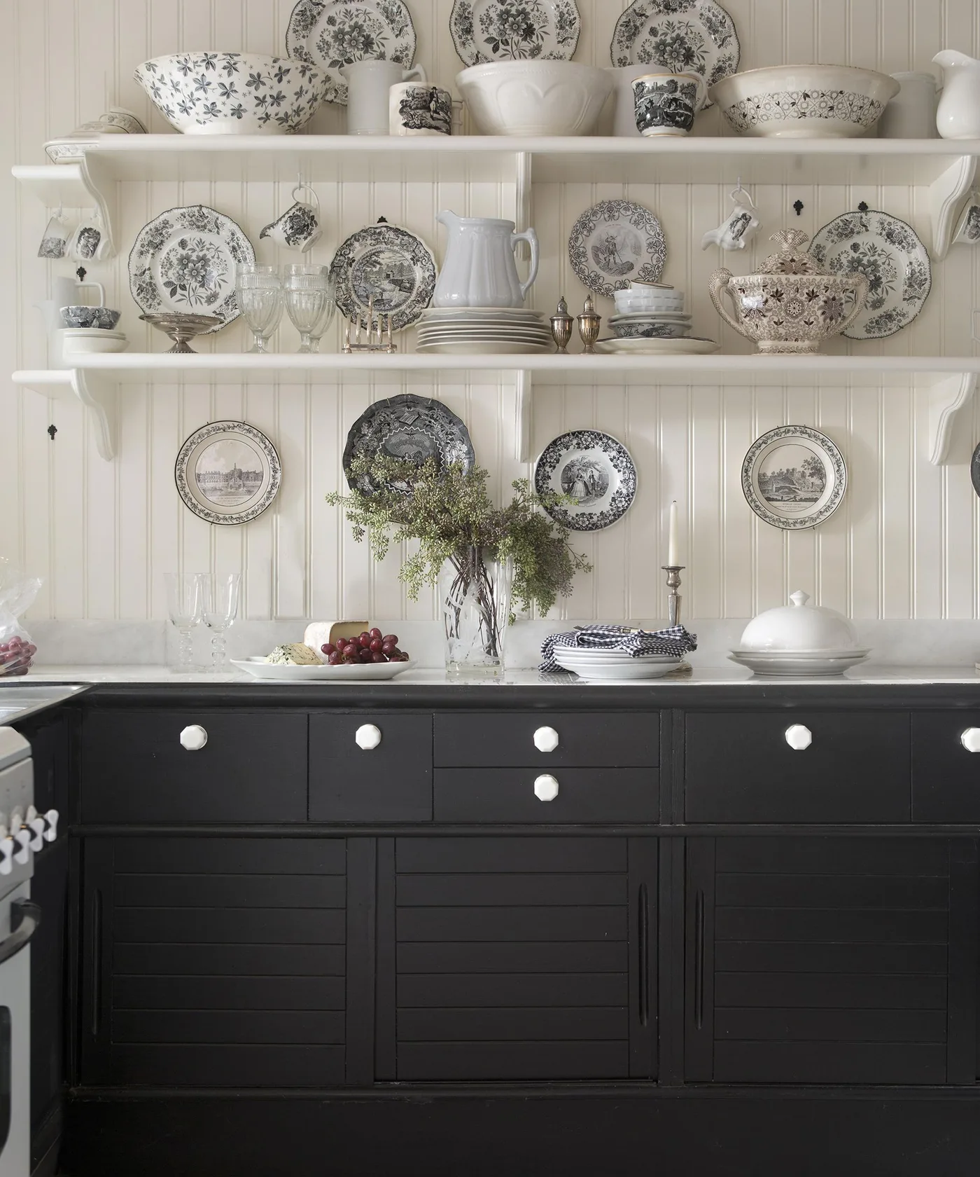 Near-black kitchen base cabinets with white round knobs, pale counter, white paneled wall, display shelves of patterned dishes, and a central vase.