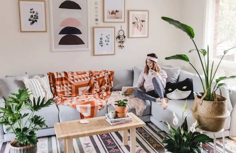 A pale gray sectional has an orange geometric throw, mixed cushions, a woven-top table, potted plants and a wall display of pale framed prints.