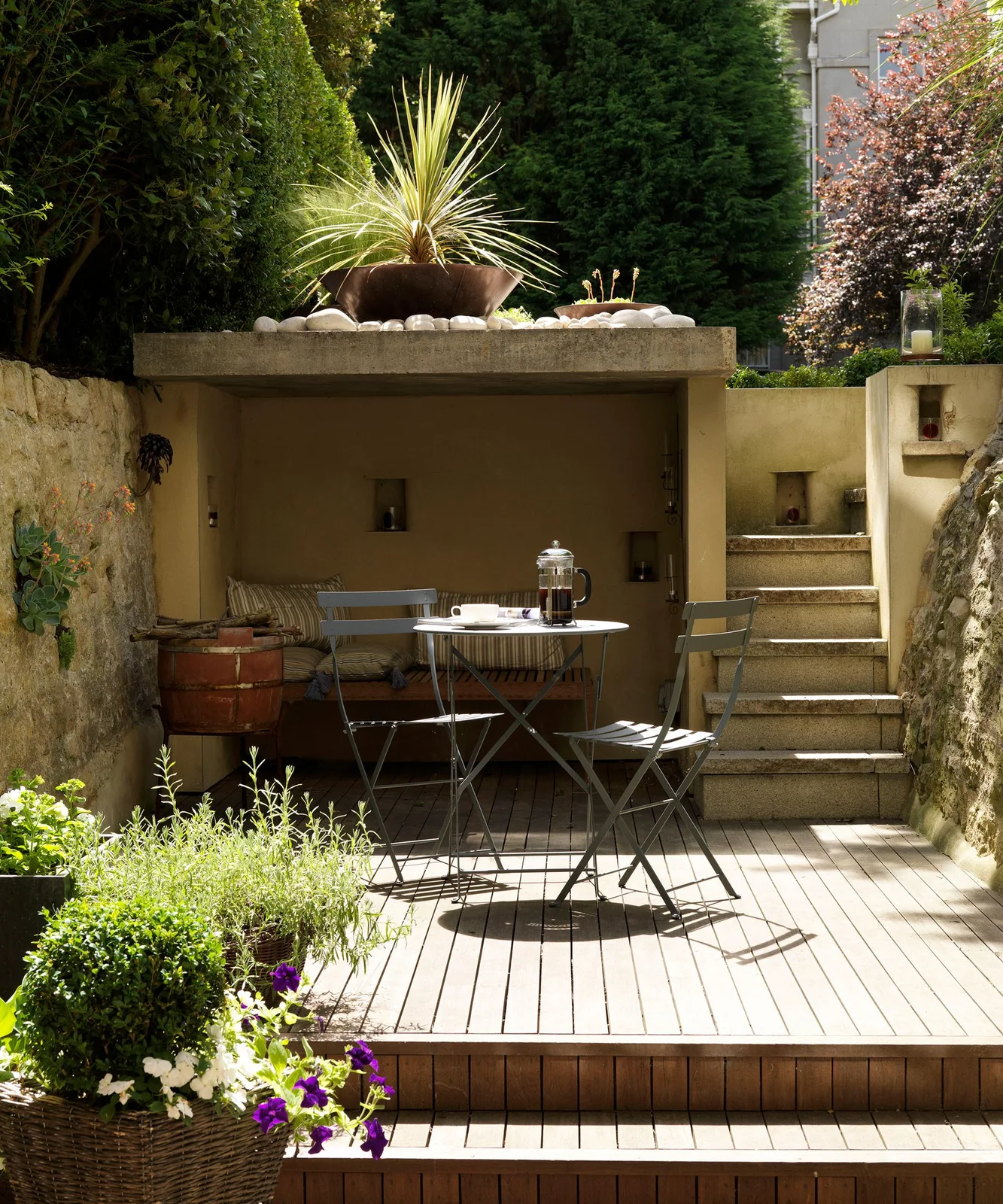 A small timber terrace with a bistro table sits below stone walls beside garden steps