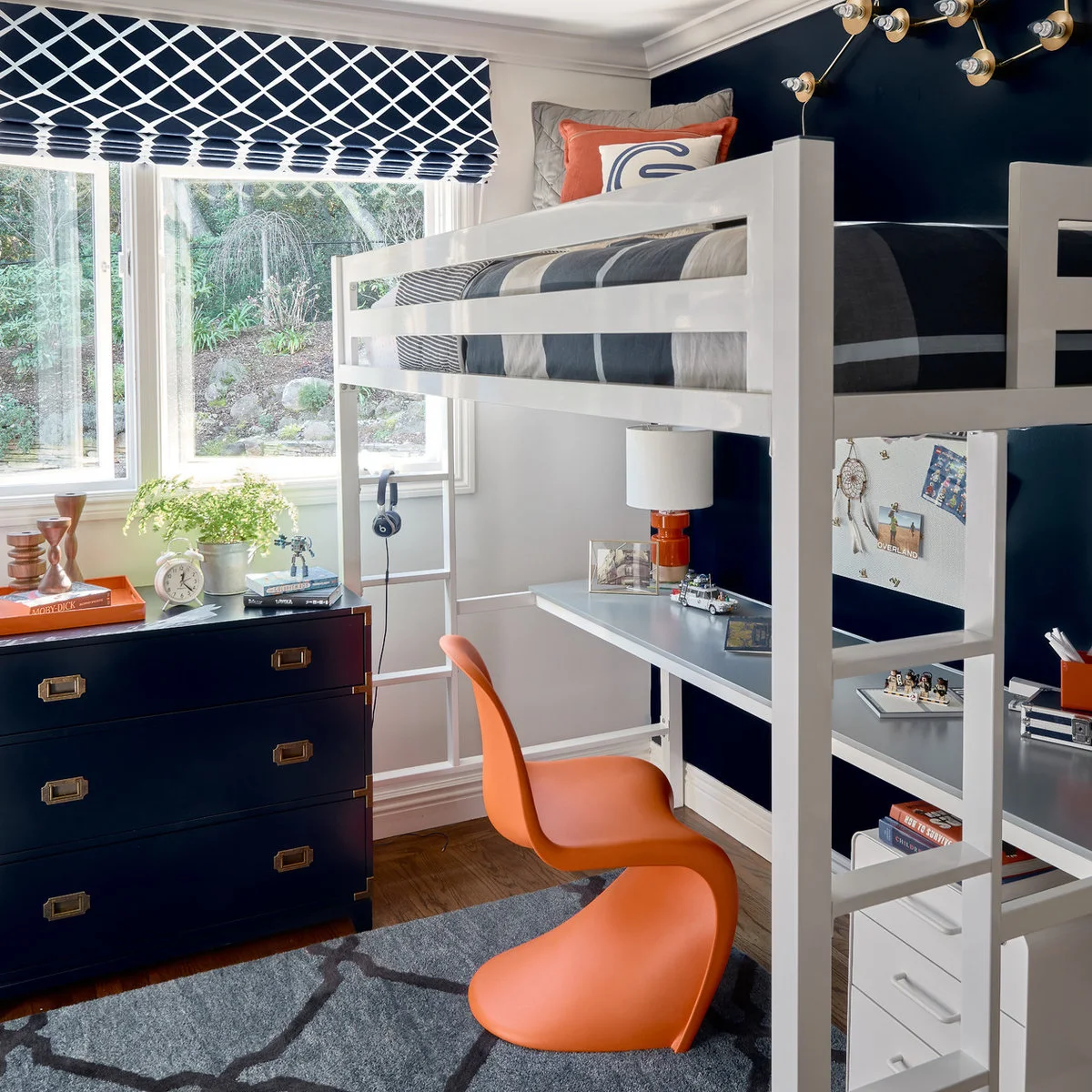 White loft bed with a desk and orange chair below, navy walls and navy-and-white window fabric.