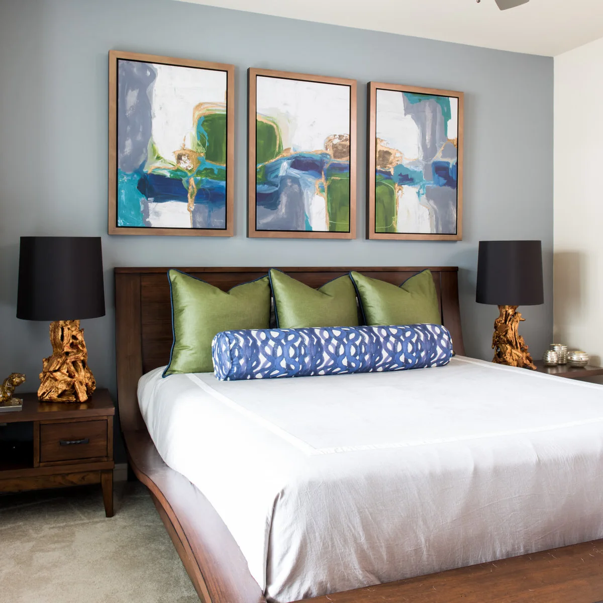 Three blue and green abstract pictures in wood frames above a wooden bed with green pillows and a blue bolster.