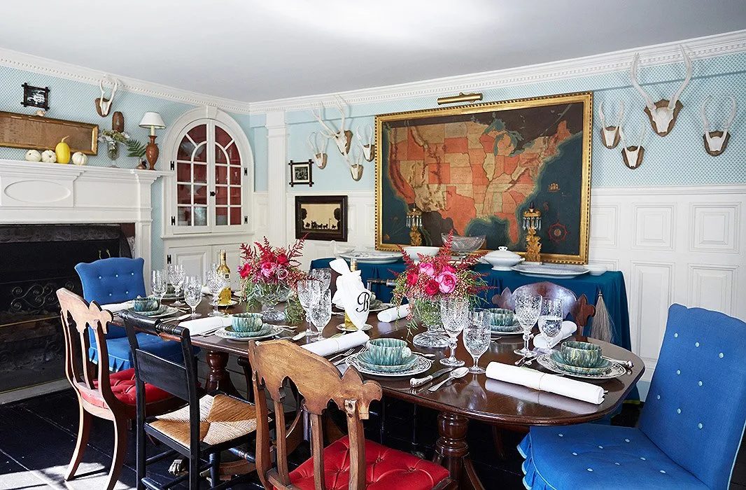 Dark oval table set with glassware and dishes among blue and wooden chairs below framed map artwork.