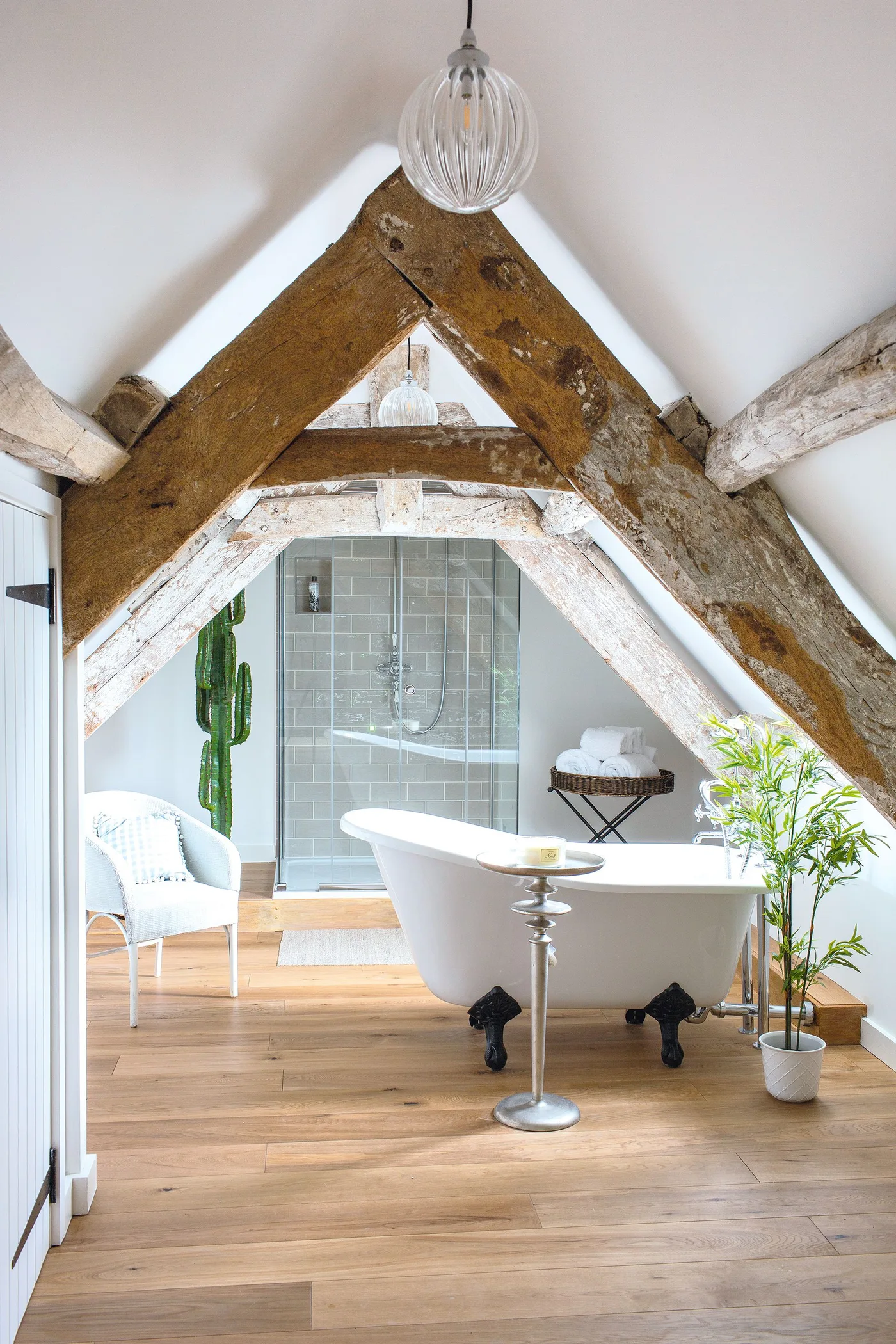 Loft bathroom with exposed sloping beams, a white claw-foot bath, wooden floor, glazed shower enclosure and plants.