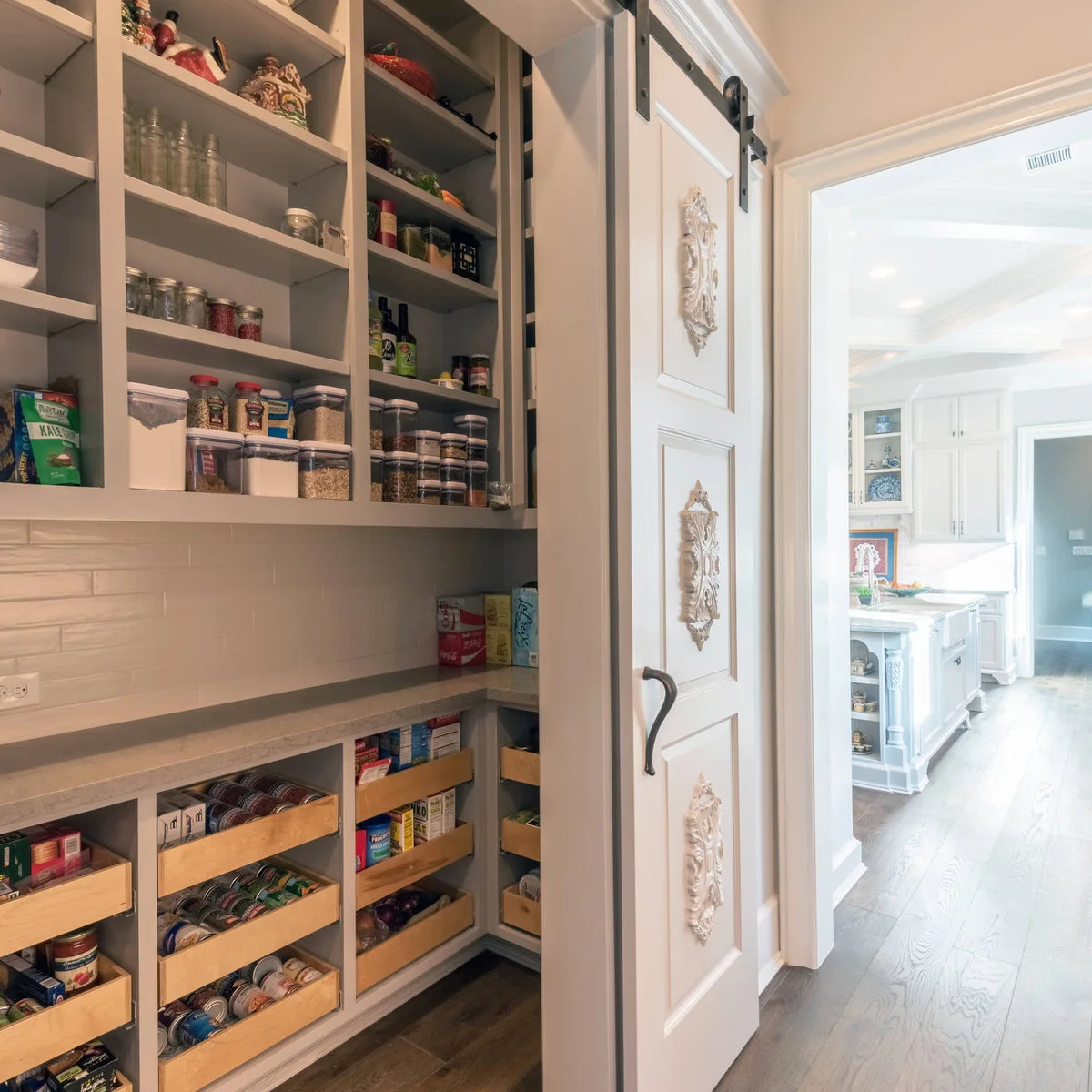 A decorative pale sliding pantry door opens onto upper food shelves, a long counter and lower wood pull-out trays.