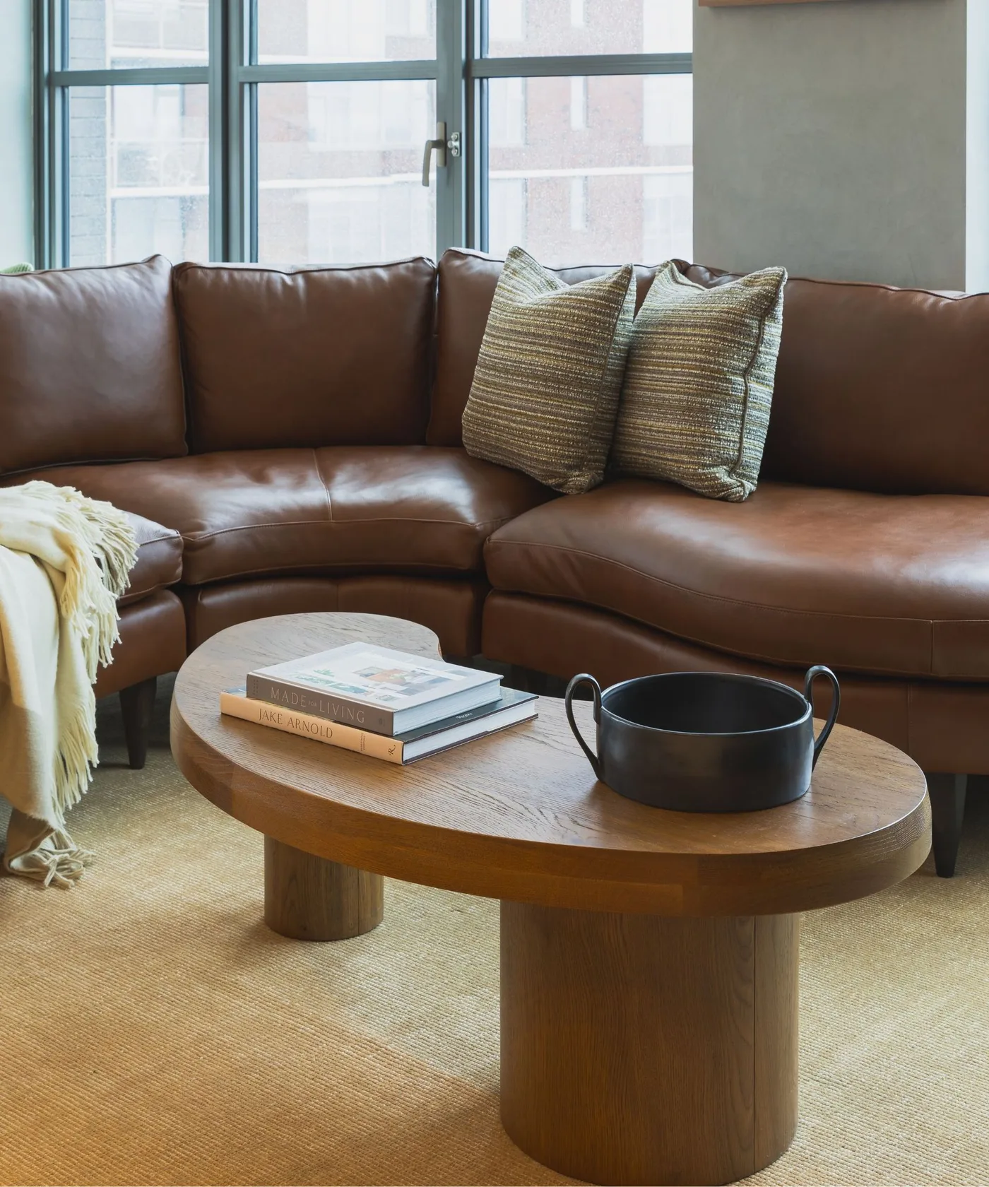 A brown leather corner couch sits beneath large windows beside two striped cushions, a rounded wooden table and pale rug
