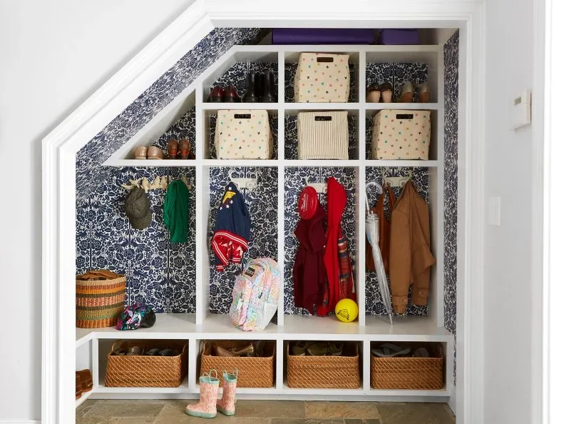 A white bench and basket bays fit below a sloping staircase, with blue floral wallpaper, coat hooks and varied upper cubbies.