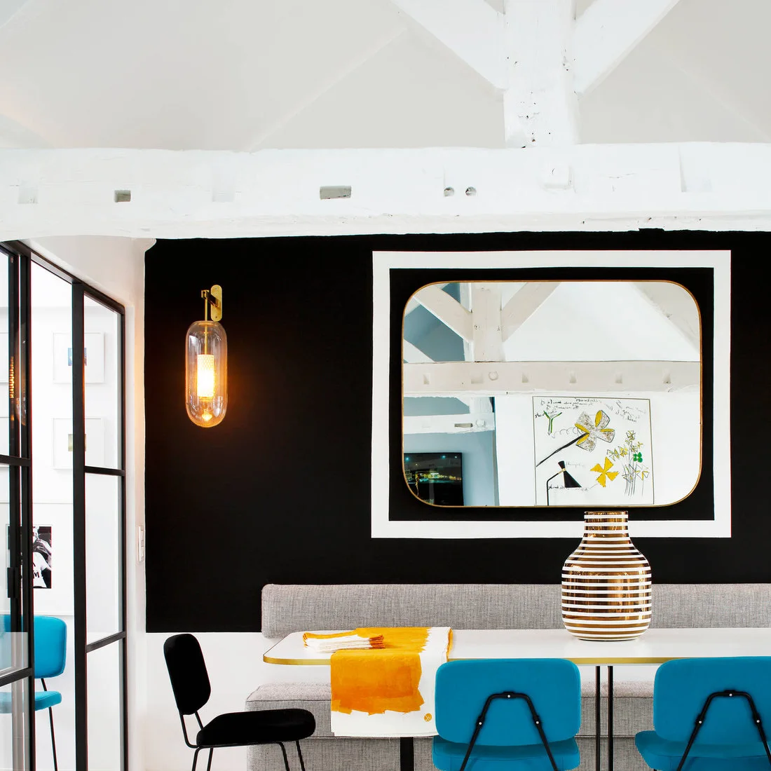 A pale dining table, gray banquette and bright blue chairs sit against a black wall with a large rounded rectangular mirror.