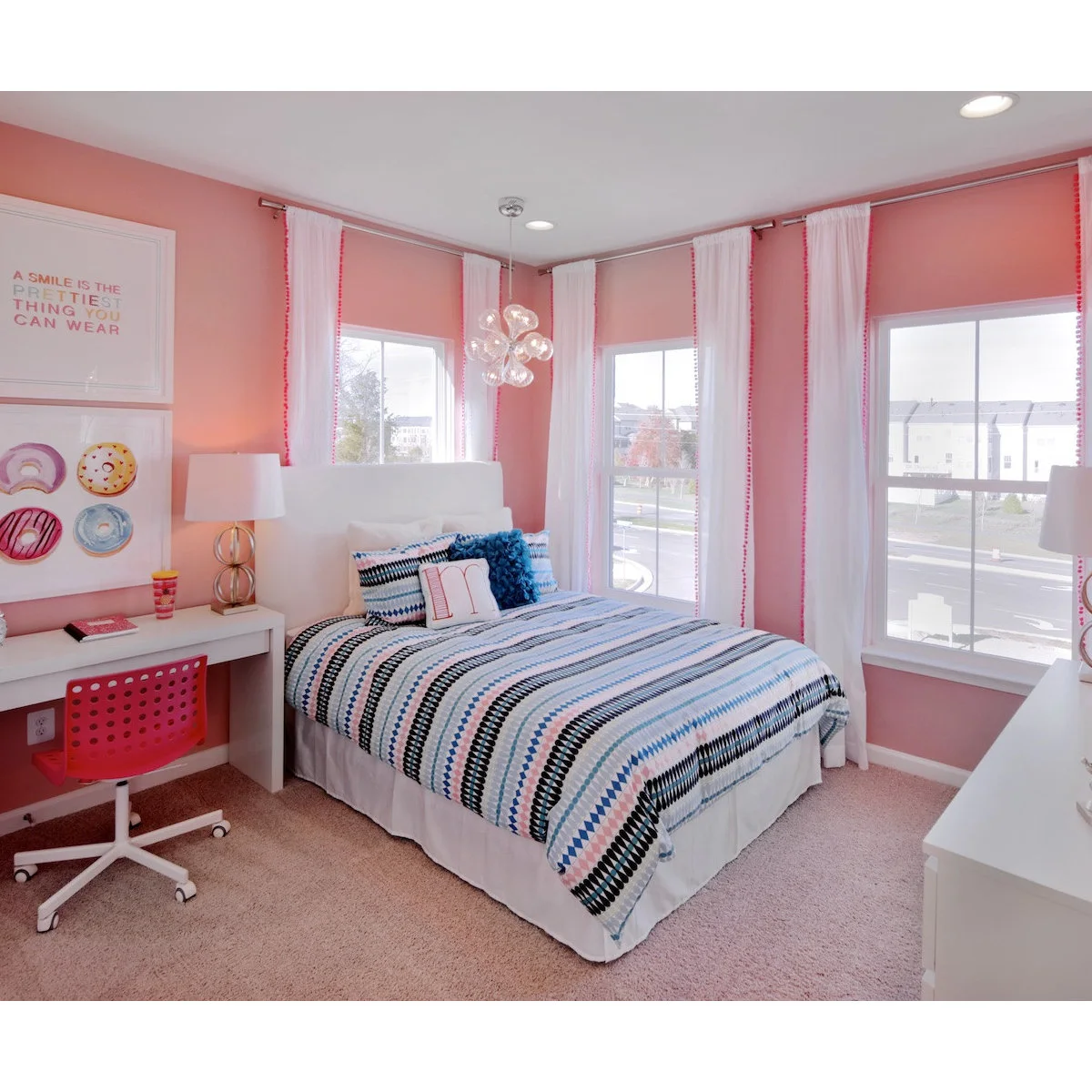 Coral-pink bedroom with tall white curtains, striped bedding and a desk beside the bed