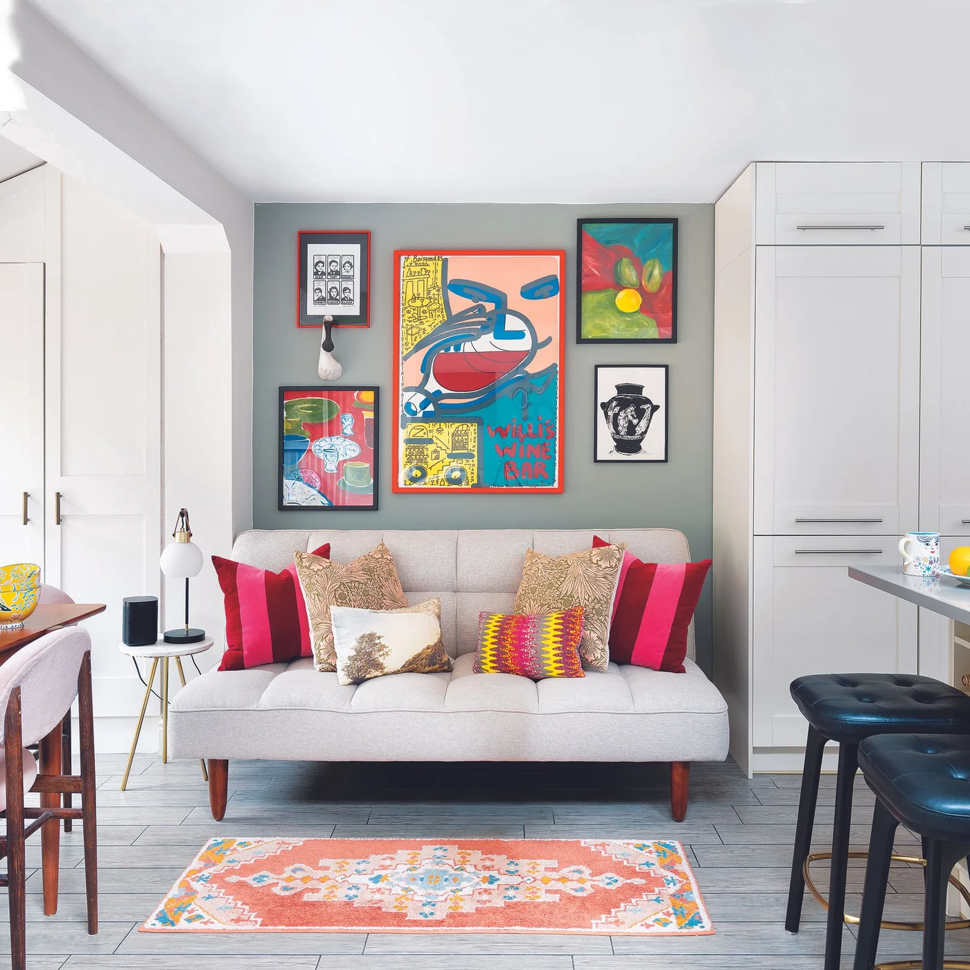 Colorful framed pictures above a pale sofa between white kitchen storage and an island with black stools.