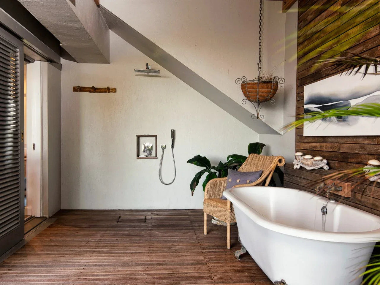A white tub beside timber wall boards, woven chair and an open shower area under a slope