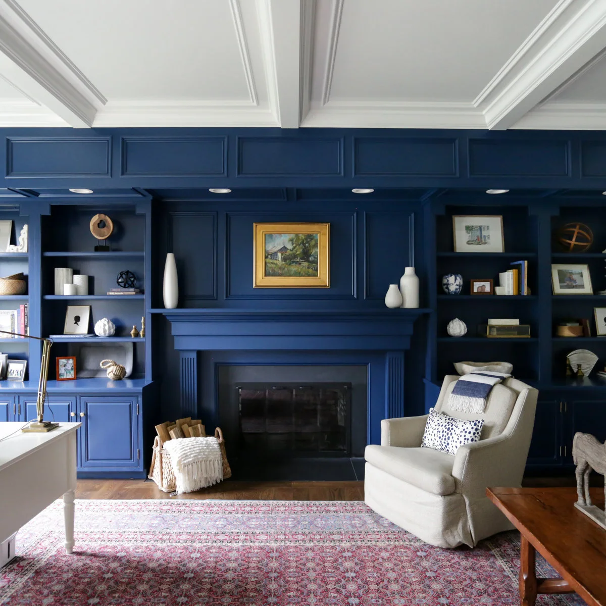 A pale reading chair and low wood table beside a white desk in front of deep blue fireplace shelving.