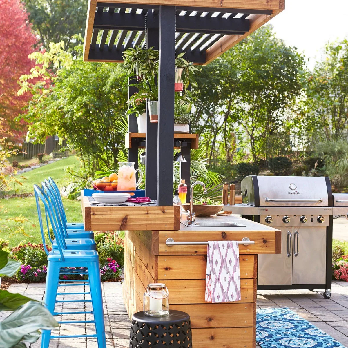 Wood-clad outdoor bar with bright blue metal stools, a small sink and plants hanging from a dark pergola post