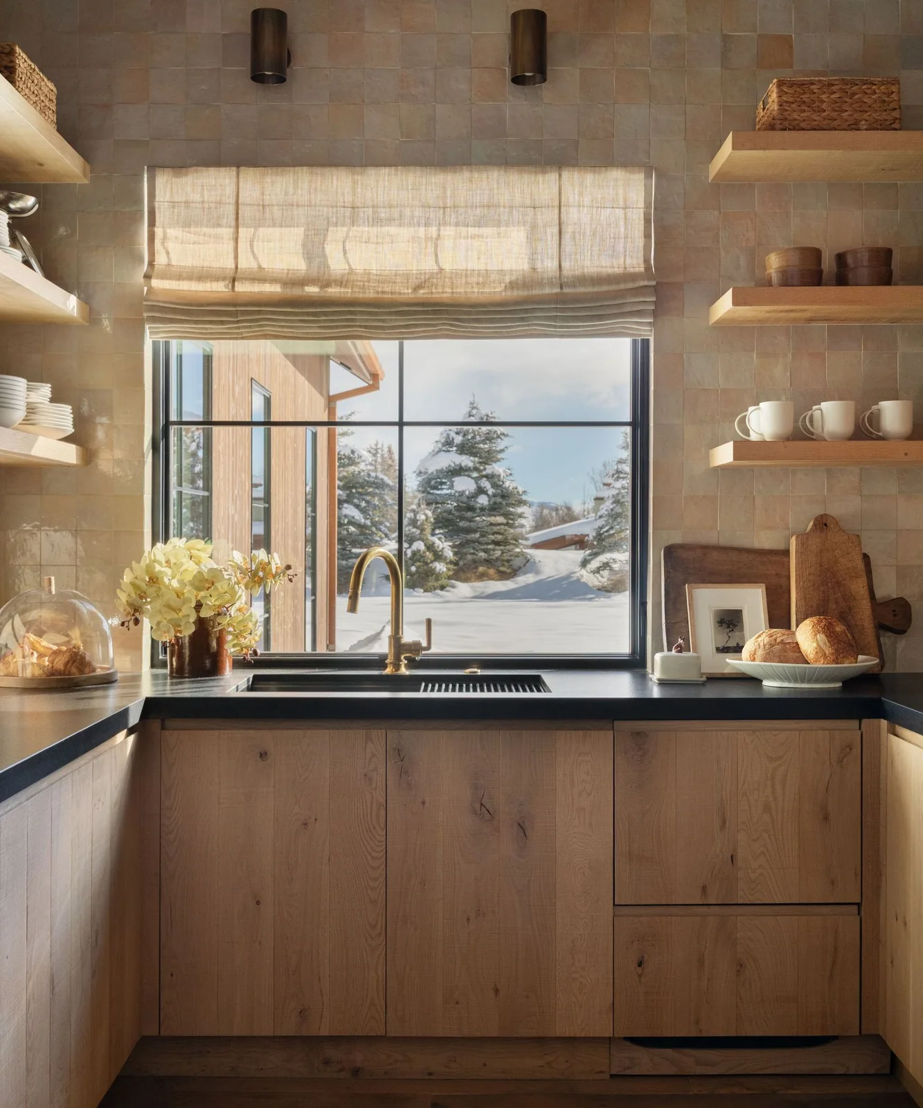 Oak pantry cabinets sit below a black counter and a snowy garden window, with pale tiles and open shelves on both sides.