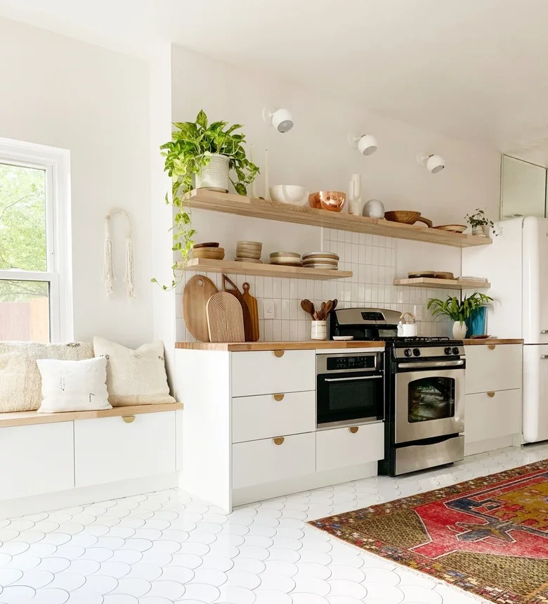 A white kitchen has flat drawers, wood counters and shelves, a cushioned window bench with storage below, a black range and a red patterned runner.