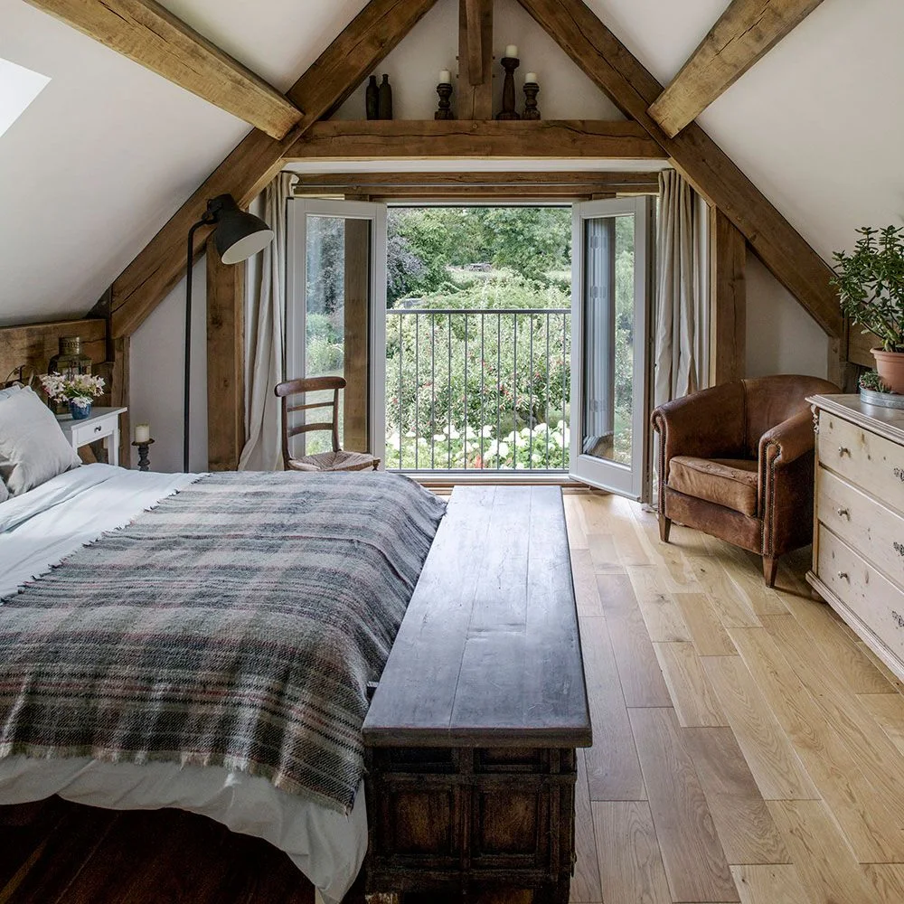 Bed and wooden bench beneath exposed attic beams with glazed doors and a brown chair at the far end.