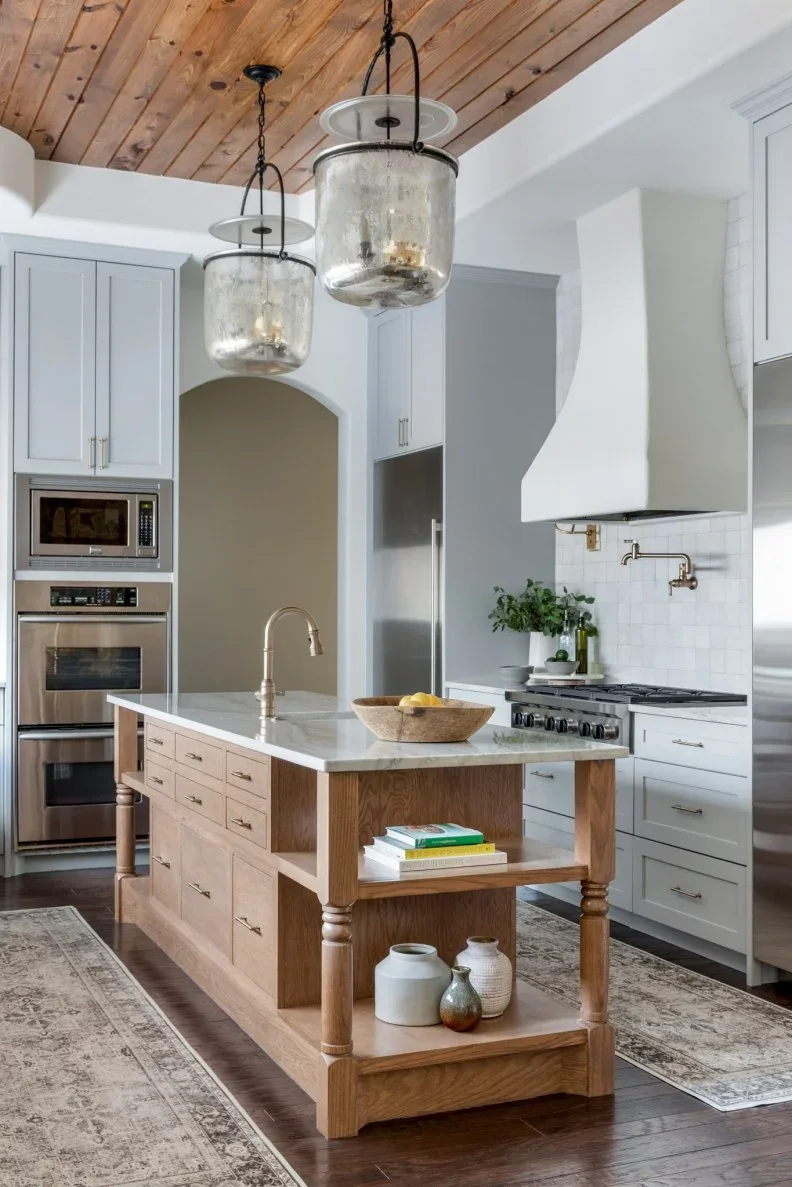 A wood island with turned legs, drawers and open end shelves stands among pale painted cabinets beneath a wood ceiling.