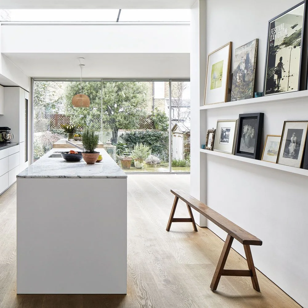 Two white picture ledges with mixed framed art above a wooden bench beside a white kitchen island.