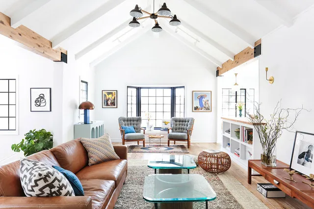 A tan leather sofa sits beside pale tables and blue cushions in a long room with a pitched white ceiling