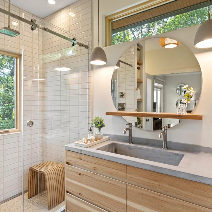 A wood vanity with a long gray basin and two taps sits beneath a round mirror and high window beside a white-tiled shower.