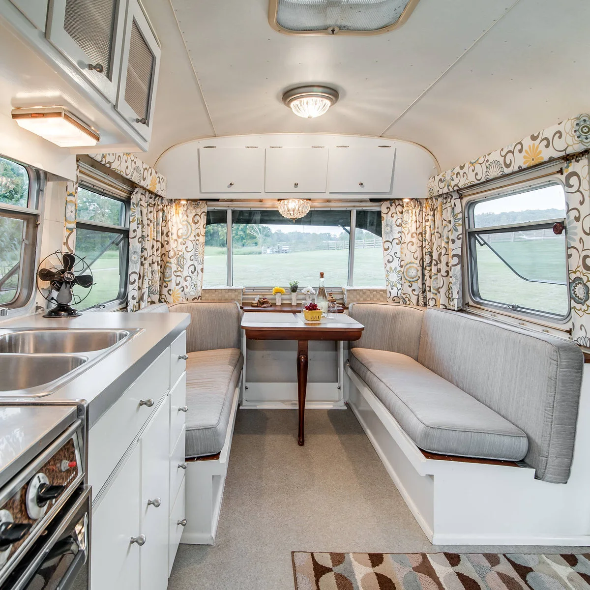 Camper dinette with two pale upholstered benches, a central table and patterned curtains around three windows.