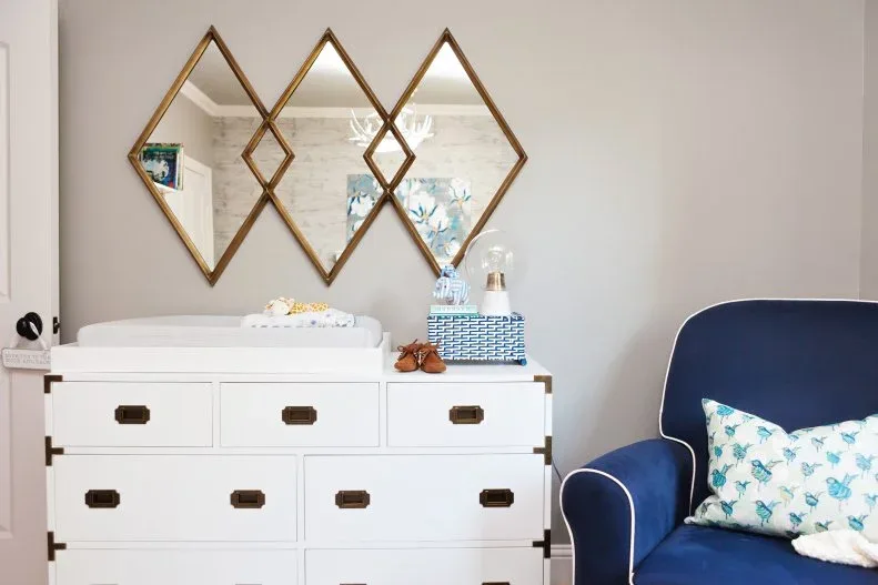 White nursery dresser with dark handles beneath diamond-shaped mirrors beside blue upholstered chair