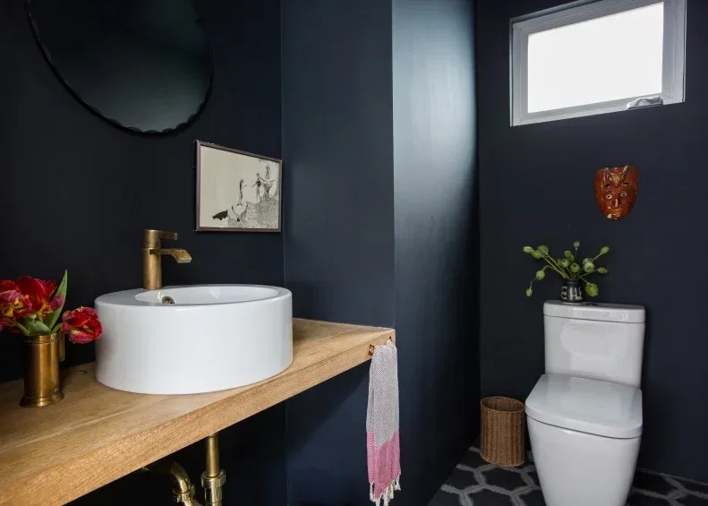 A white round vessel basin sits on a plain wood counter beneath a dark round mirror beside deep blue walls, a small window and a white toilet