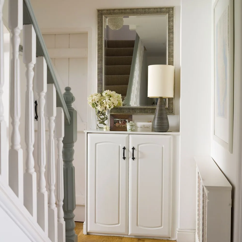 Pale hallway with a white paneled cupboard, tall framed mirror, shaded lamp and gray-green stair trim.