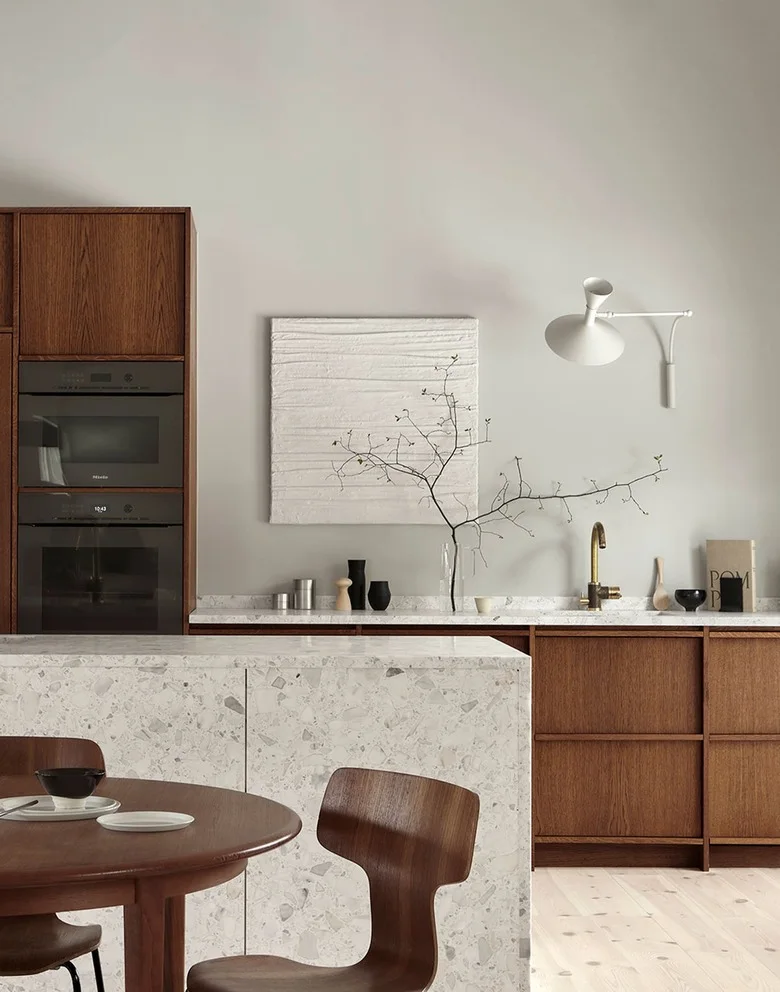 A kitchen has medium brown wood cabinetry, pale walls, a light patterned island surface, a round wood table and a white adjustable wall light.