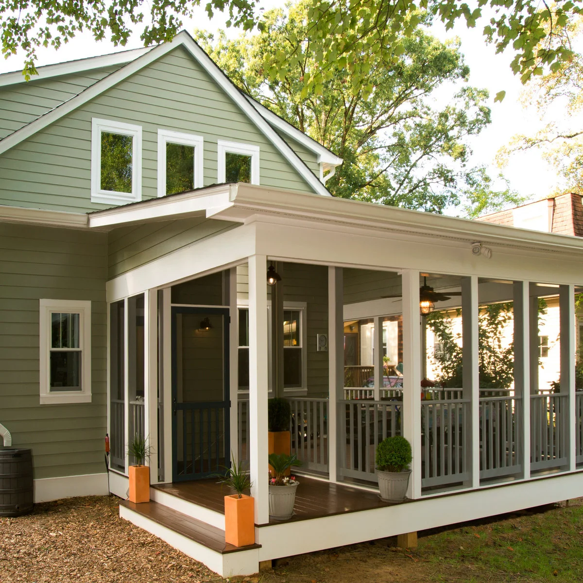 A pale green house has a screened side porch with white framing, a dark screen door, wood-toned flooring and small potted plants.
