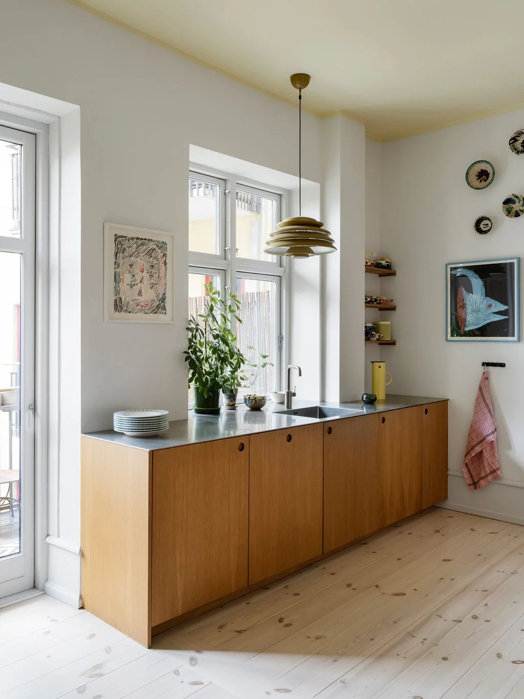 A simple wood kitchen run with circular pulls sits beneath tall windows, with a slim metal counter, pale floorboards, a layered pendant and small wall artwork.