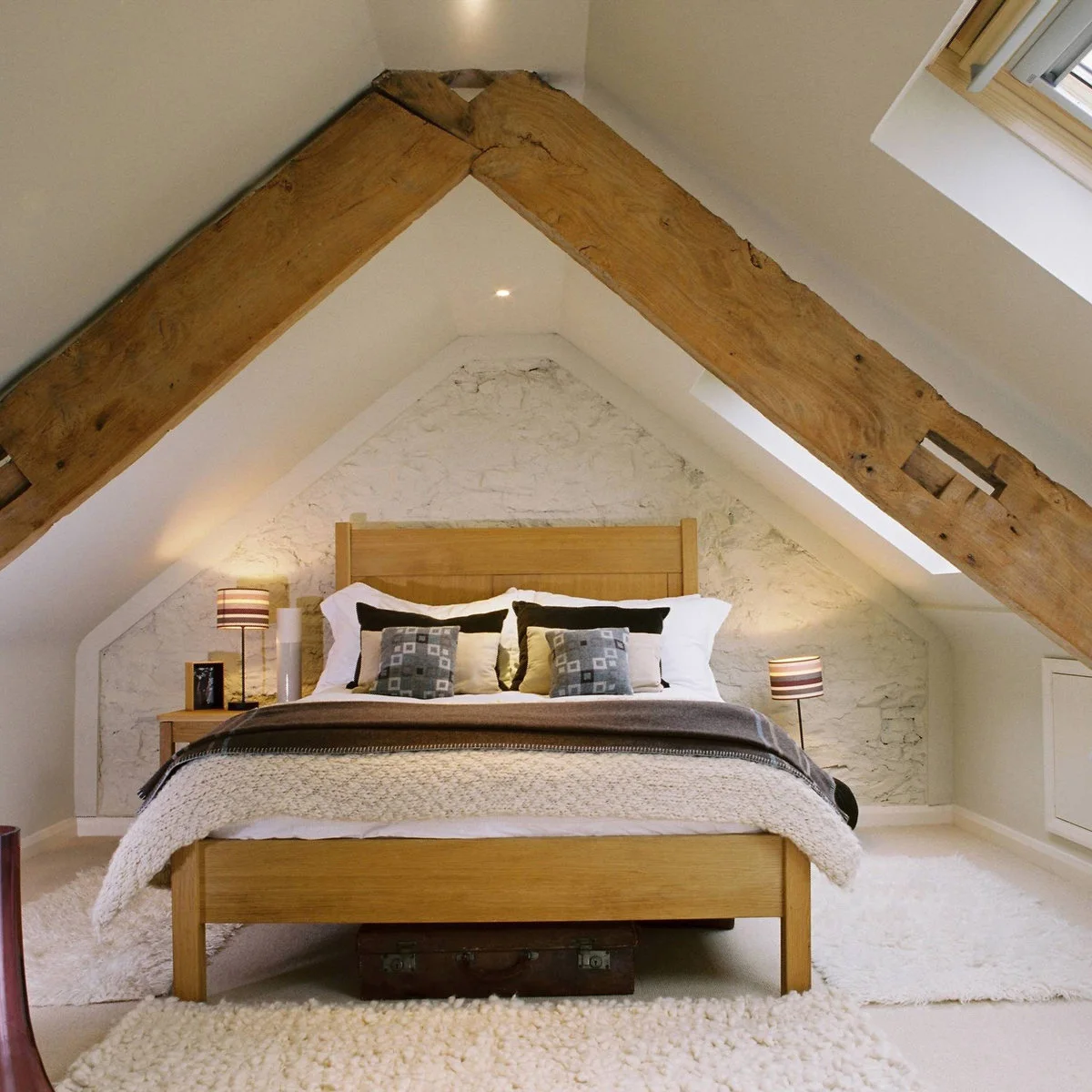 Attic bedroom with large exposed beams, simple wood-colored bed and layered pale rugs