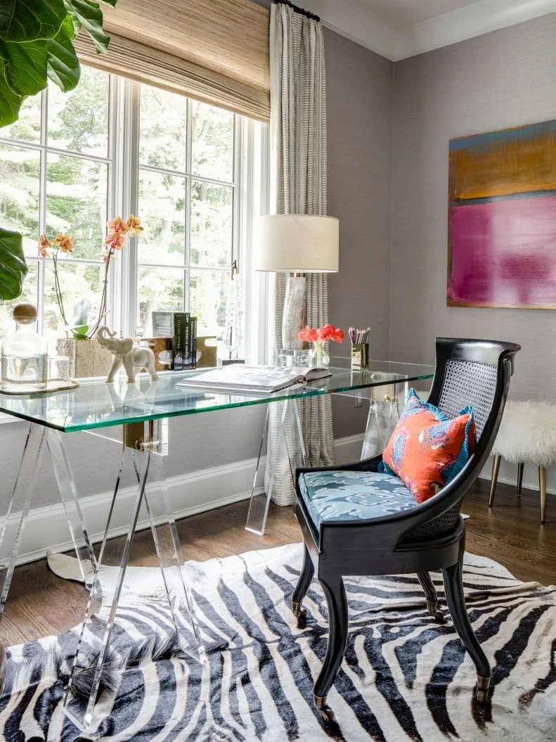 A glass-topped desk with clear supports stands by a window beside a dark cane-backed chair, patterned rug and colorful artwork.
