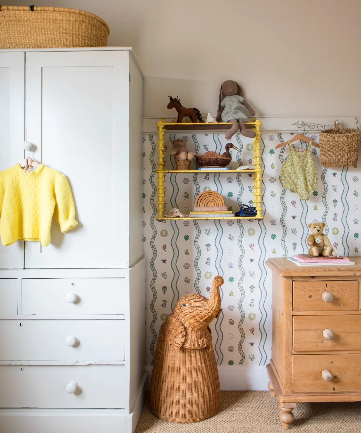 Tall white wardrobe beside warm wooden dresser, finer lower-wall wallpaper, small yellow shelf and baskets.