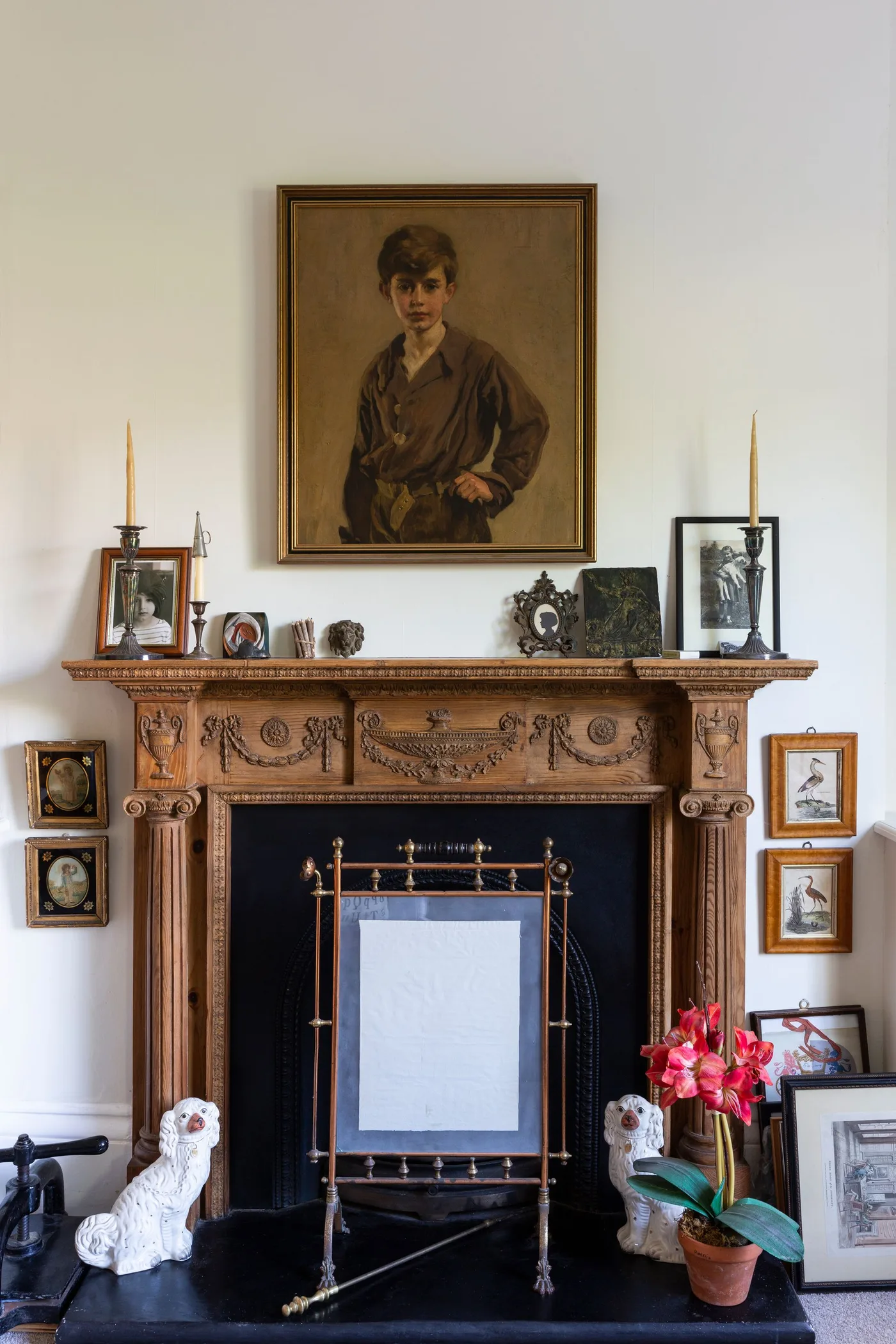 Carved wooden fireplace with portrait and smaller pictures on mantel, framed display in opening and pale walls.