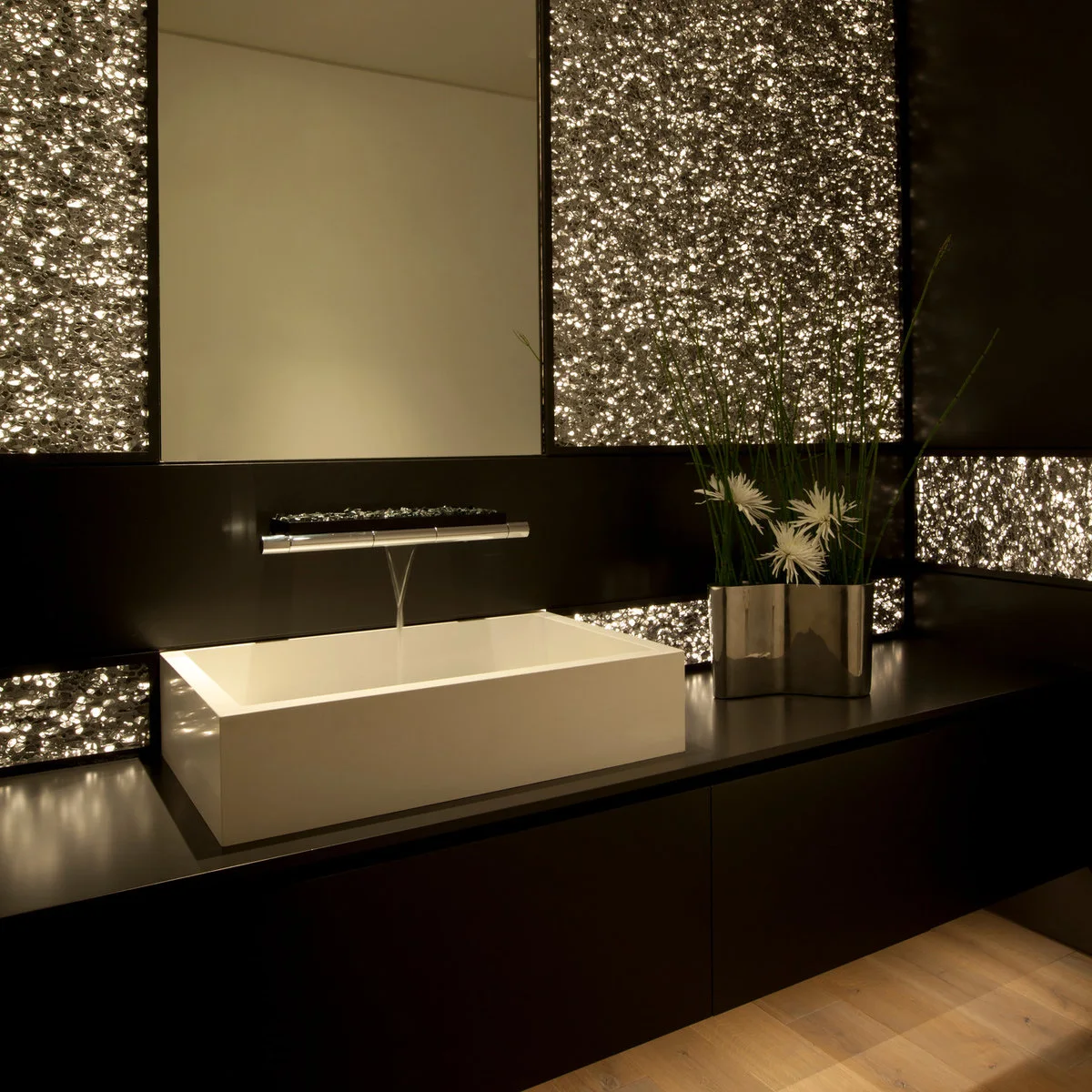A white rectangular vessel basin sits on a dark counter below a wide wall-mounted faucet, with illuminated textured panels behind it.