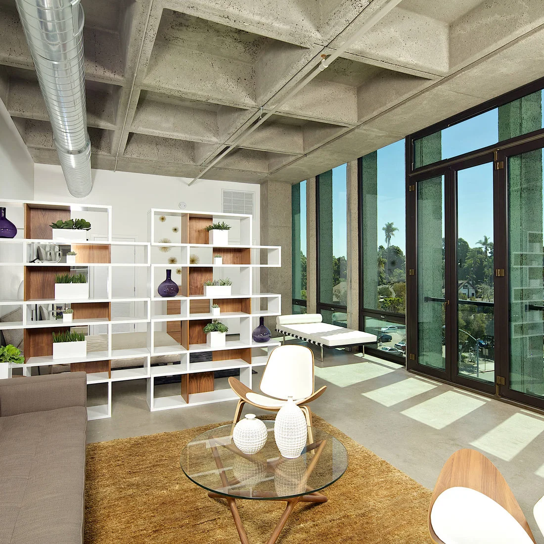 A gray sofa and curved wooden chairs beside an open white divider under a concrete ceiling