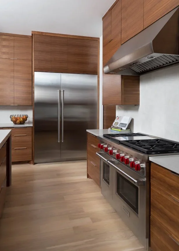 Flat walnut cupboards with horizontal grain surround a stainless refrigerator, range and hood beside pale counters.